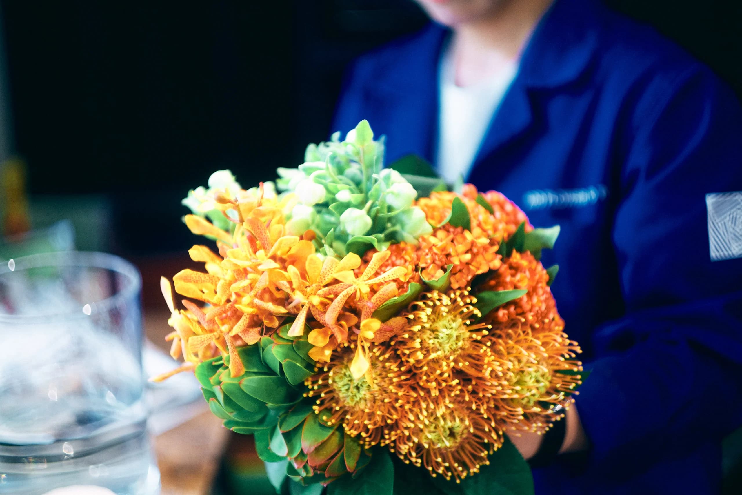 オレンジのピンクッションの花束を抱える、青いユニフォームのフローリスト